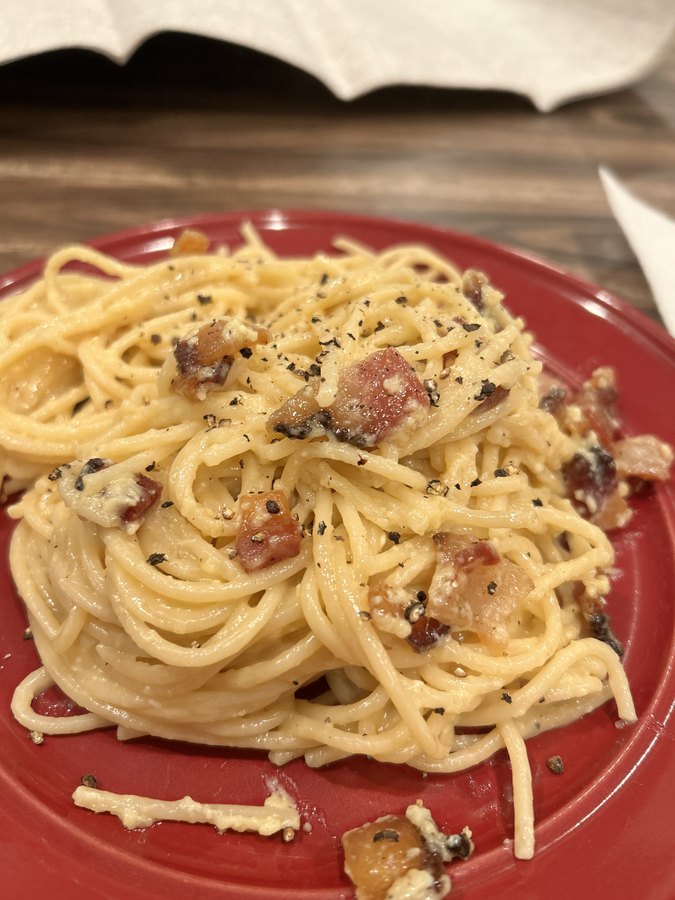 Spaghetti carbonara on a red plate, glossy with egg and pecorino, cracked black pepper on top